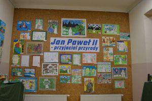Brązowa ściana z pracami dzieci i w centrum napis na białym tle niebieskie litery „Jan Paweł II przyjaciel przyrody”.