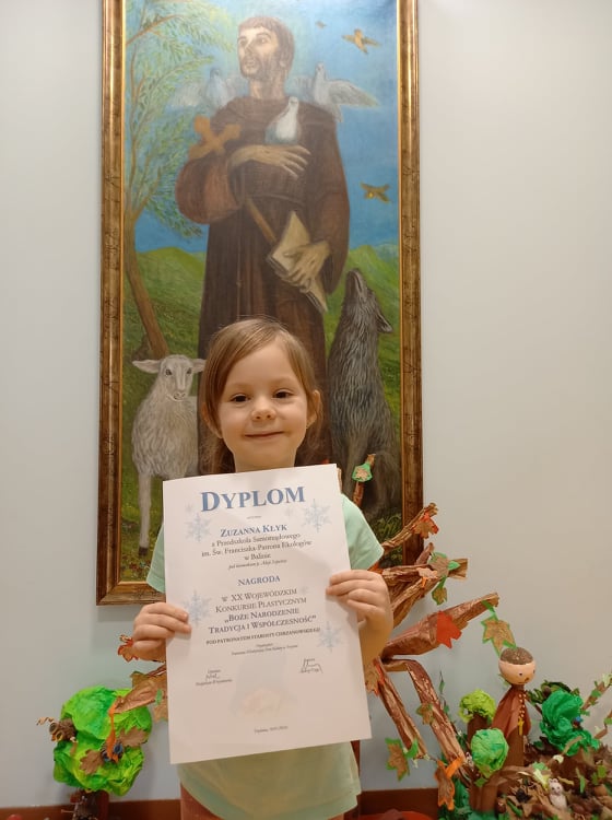 Na zdjęciu dziewczynka stoi przed obrazem świętego Franciszka, trzymając w dłoniach dyplom.