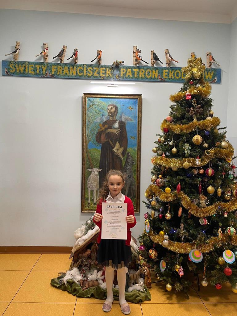Dziewczynka stoi z dyplomem przed obrazem św. Franciszka.